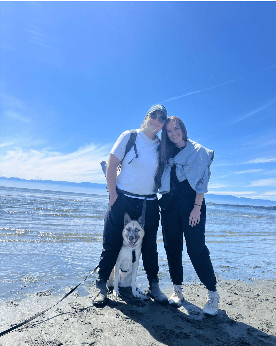 Two women at the beach with their husky dog between their legs smiling.