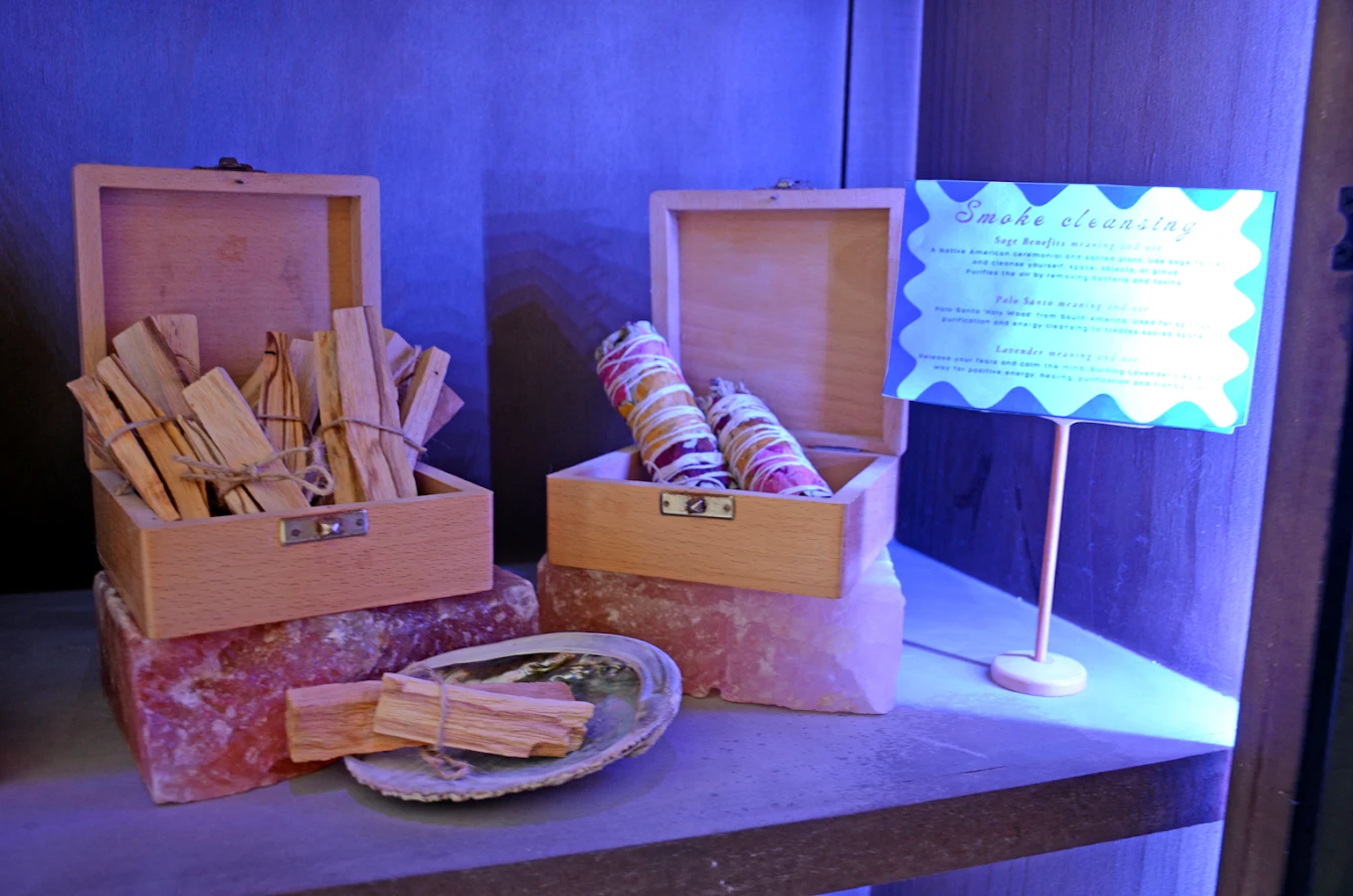 Wooden boxes with sage bundles and palo santo sticks on salt blocks, beside a sign explaining smoke cleansing benefits.