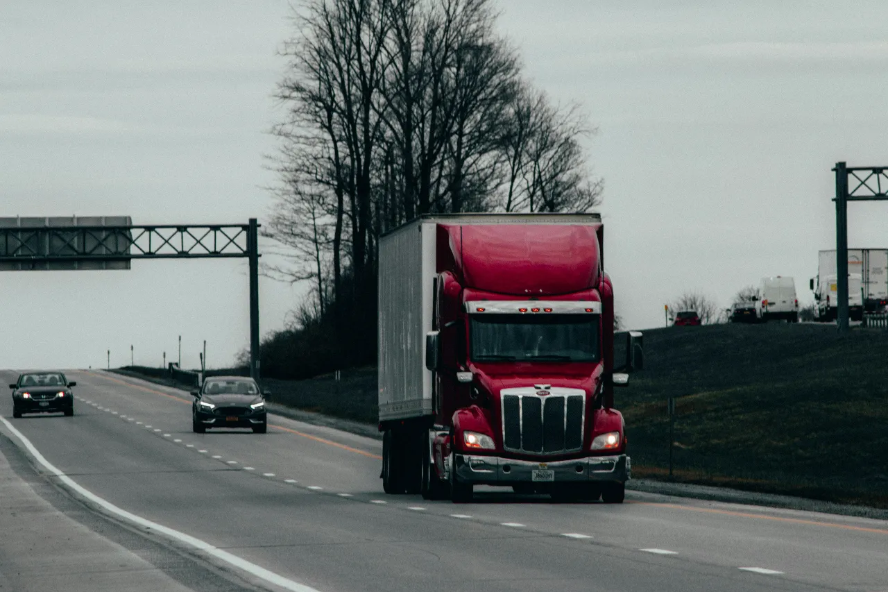 Red tractor-trailer driving along a multi-lane highway under cloudy skies, with light traffic, roadside trees, and overhead road signs visible.