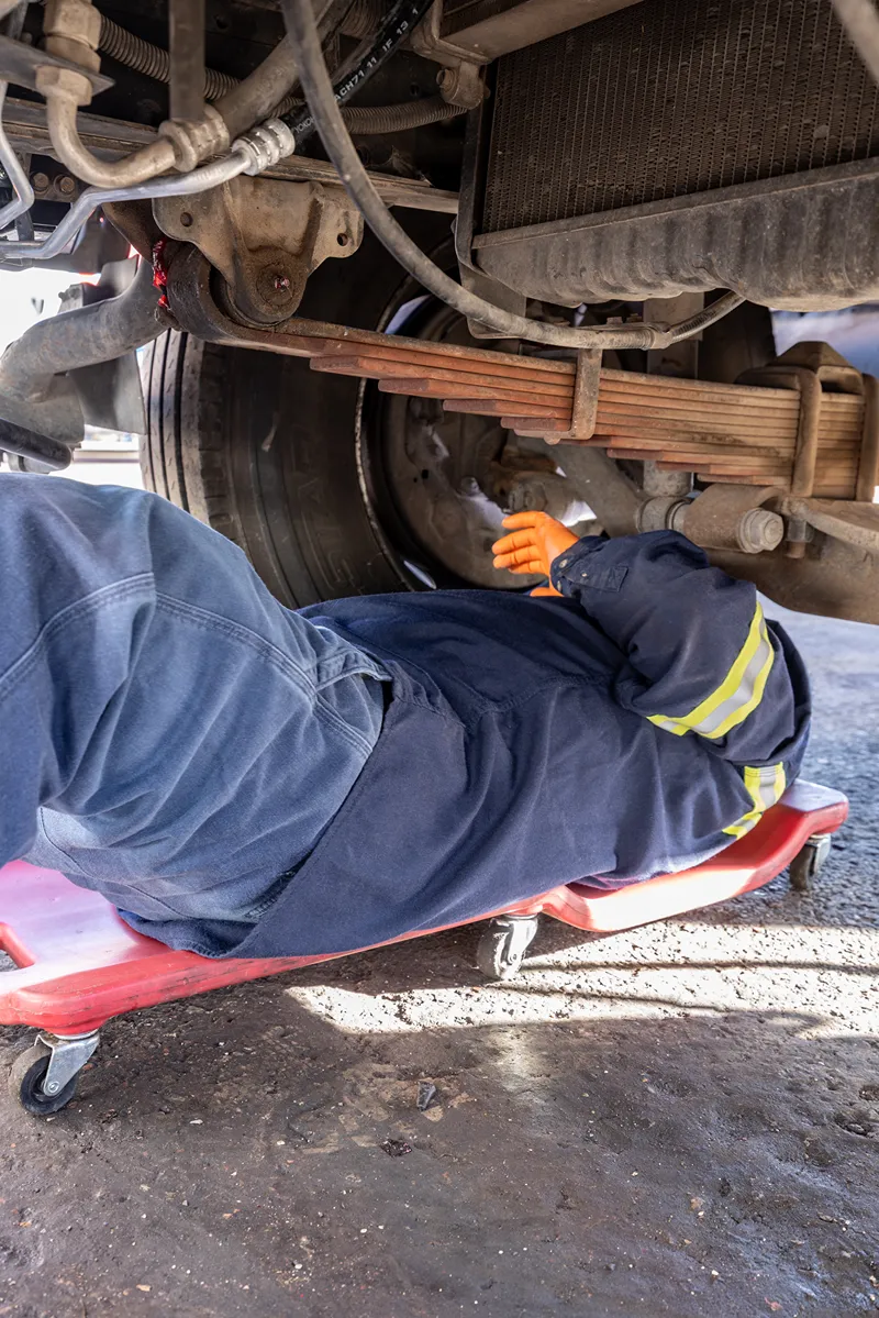 Technician inspects a heavy-duty truck suspension while working beneath the vehicle on a mechanic's creeper.