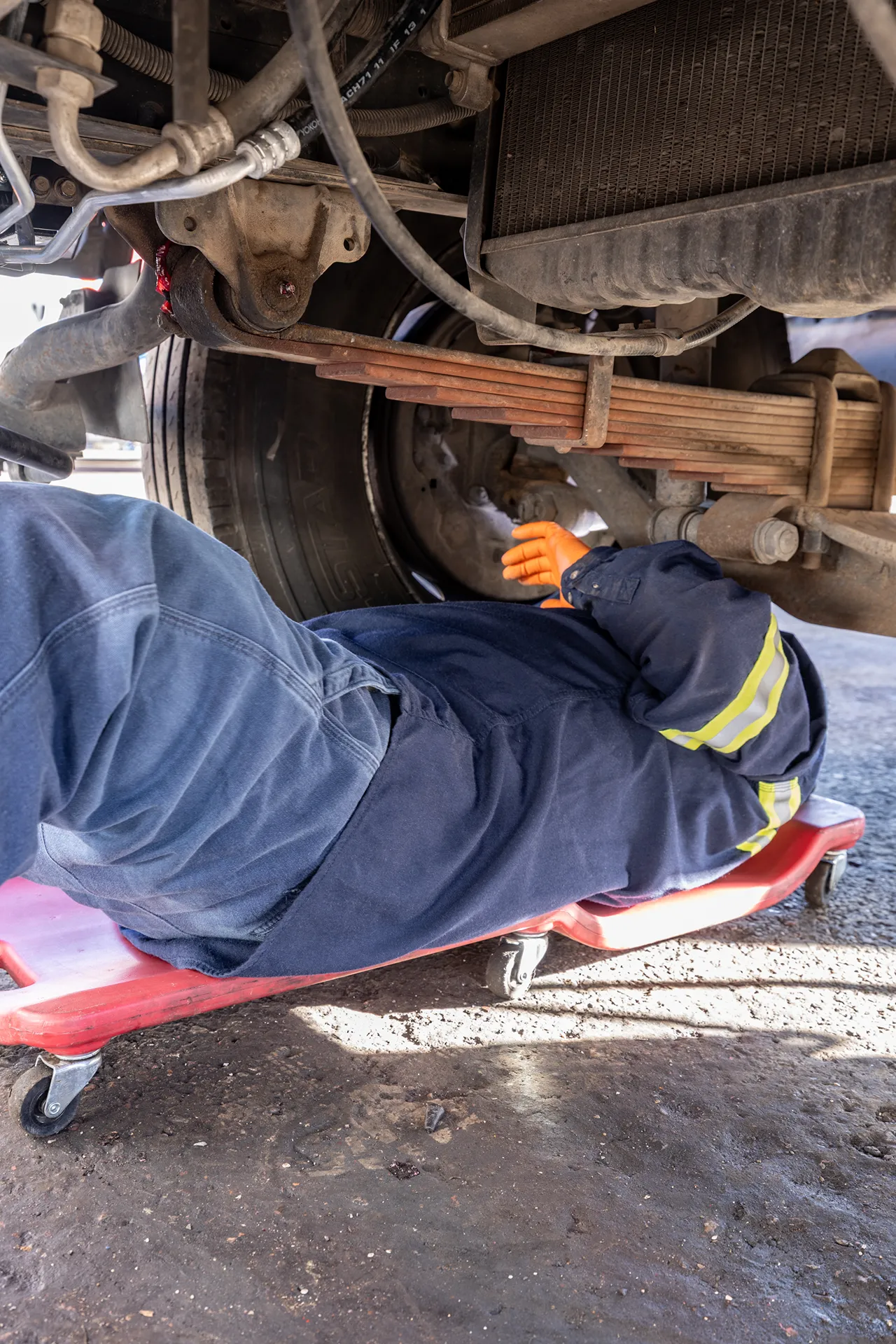 Technician inspects a heavy-duty truck suspension while working beneath the vehicle on a mechanic's creeper.