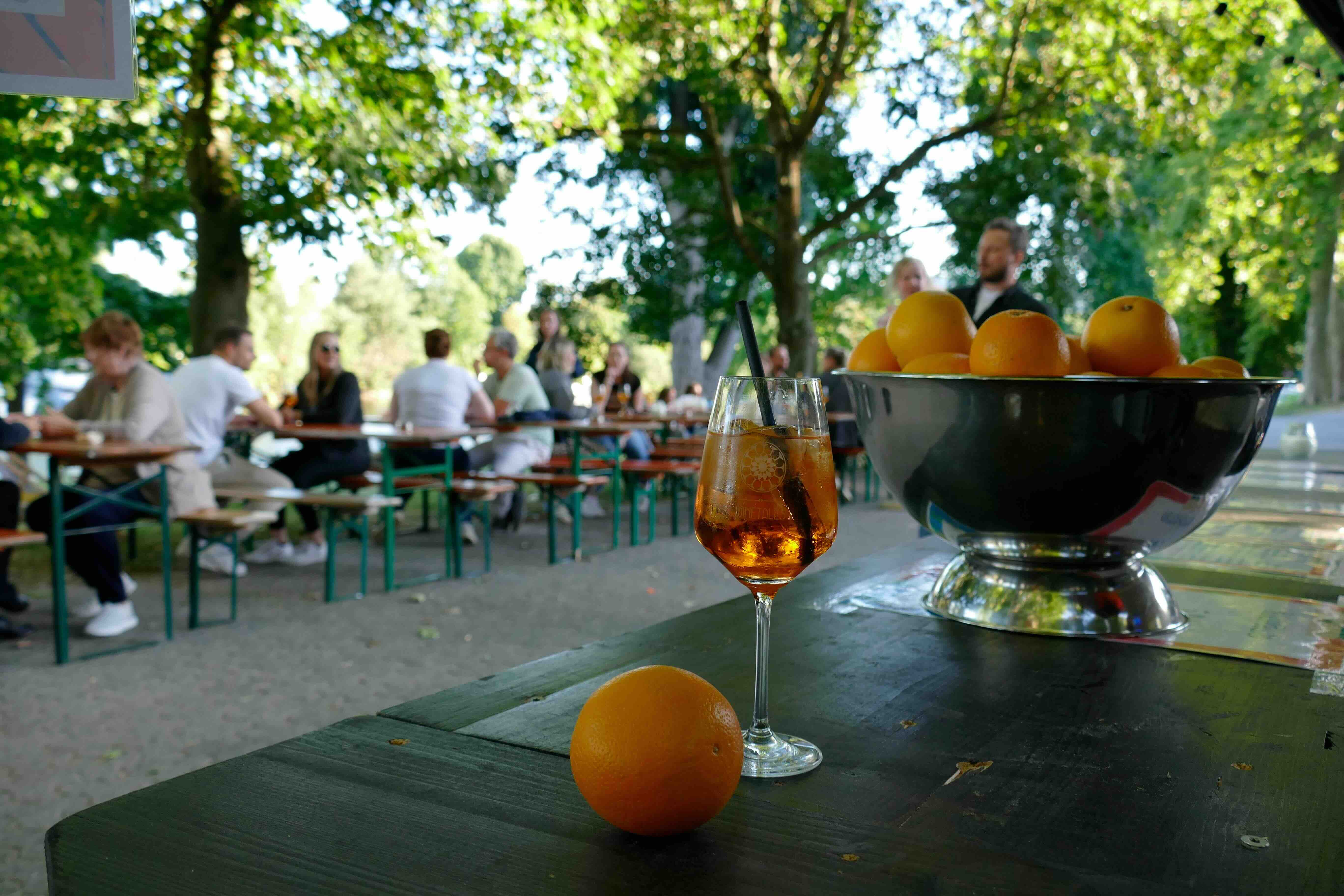 Auf einem Tisch stehen ein Weinglas und eine Orange. Im Hintergrund sieht man Menschen, die an Bierbänken sitzen.