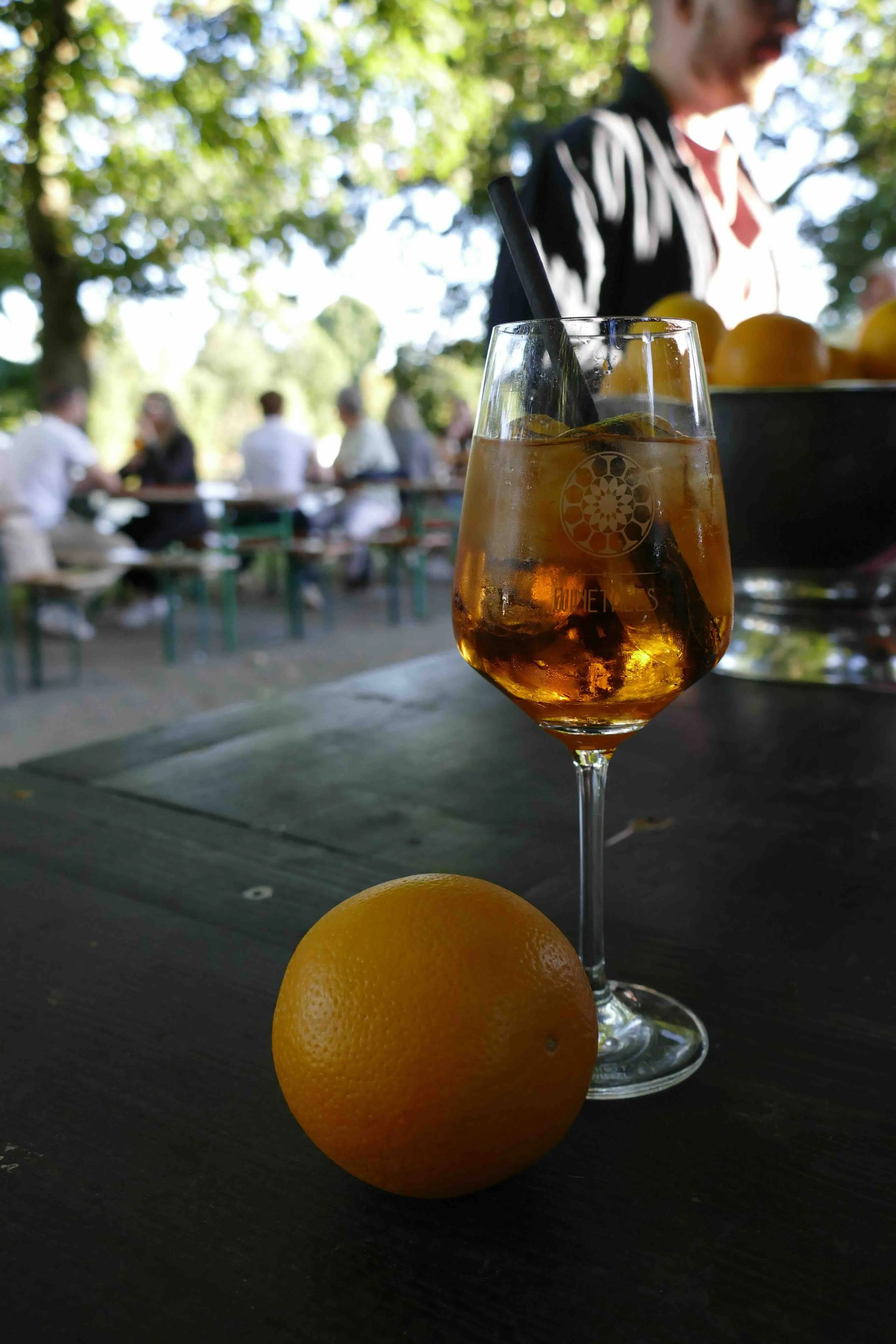 Auf einem Tisch stehen ein Weinglas und eine Orange. Im Hintergrund sieht man Menschen, die an Bierbänken sitzen.