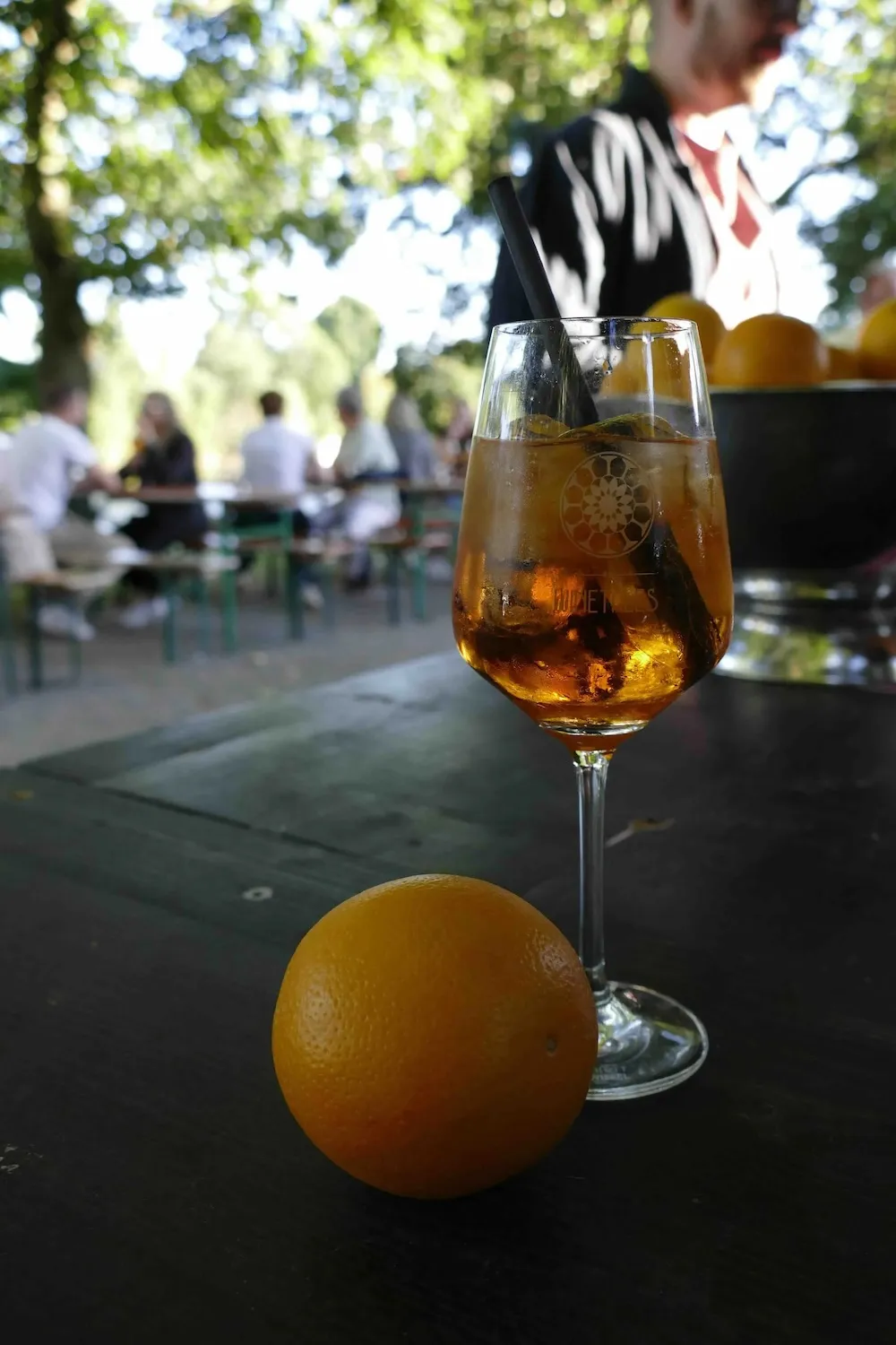 Auf einem Tisch stehen ein Weinglas und eine Orange. Im Hintergrund sieht man Menschen, die an Bierbänken sitzen.