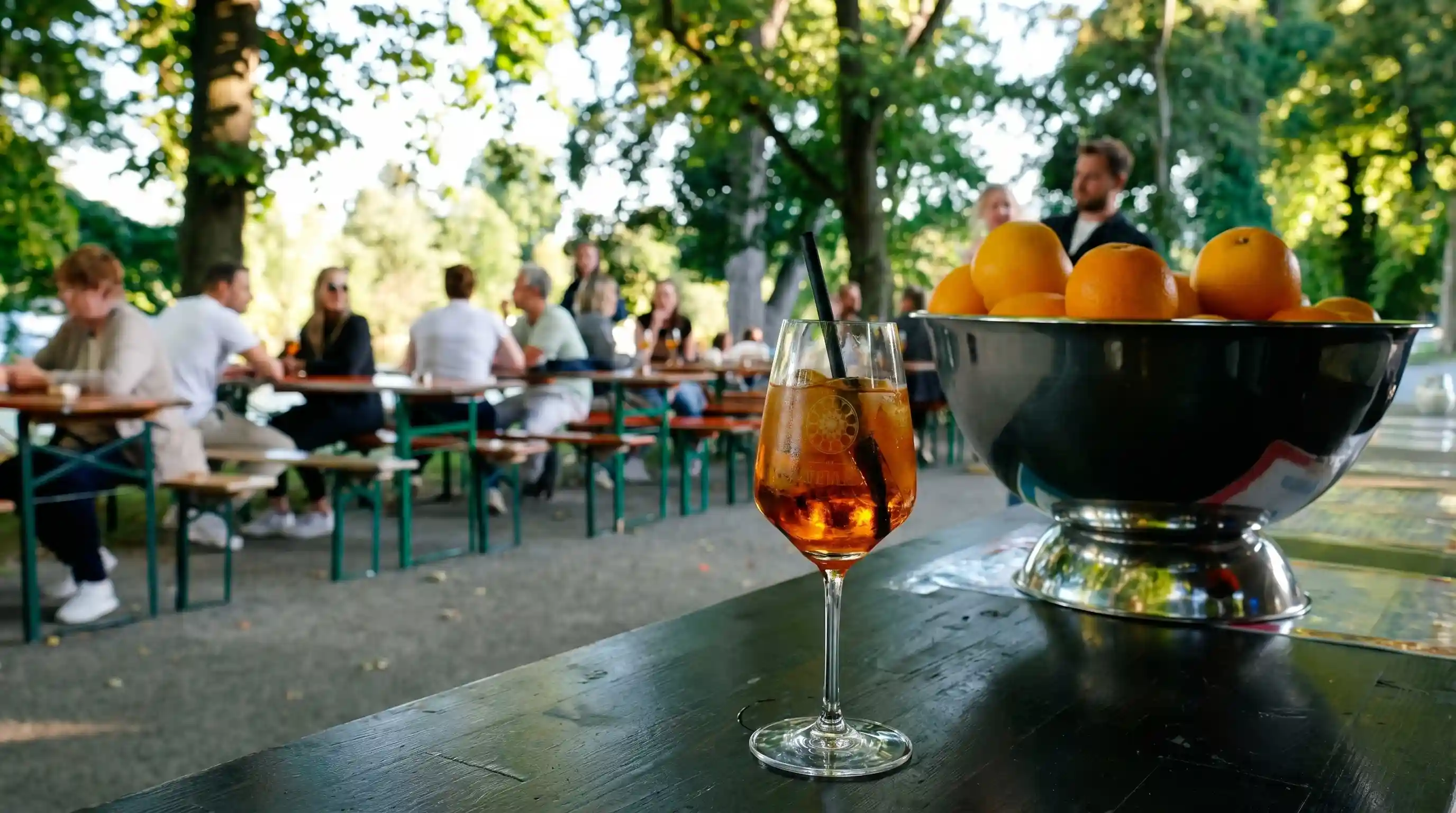 Auf einem Tisch stehen ein Weinglas und eine Orange. Im Hintergrund sieht man Menschen, die an Bierbänken sitzen.