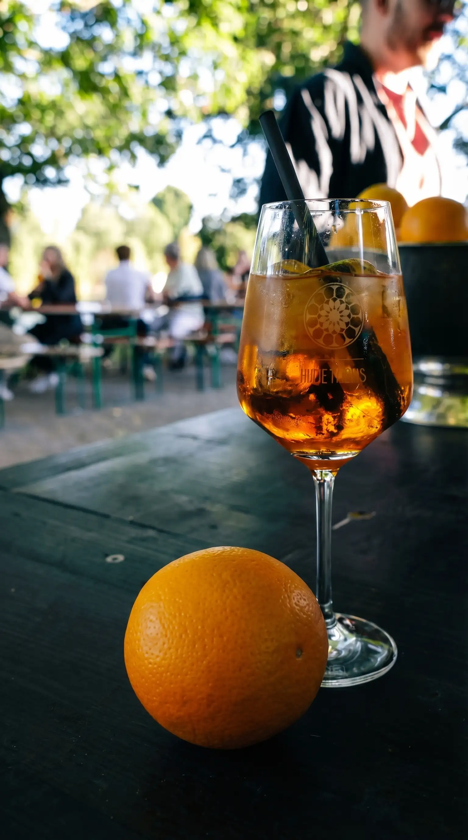 Auf einem Tisch stehen ein Weinglas und eine Orange. Im Hintergrund sieht man Menschen, die an Bierbänken sitzen.
