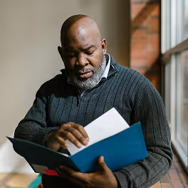 Middle-aged man with beard reading documents inside a blue folder by a window.