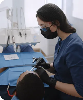 Dentist wearing protective goggles, mask, and gloves examining patient's mouth with dental instruments.