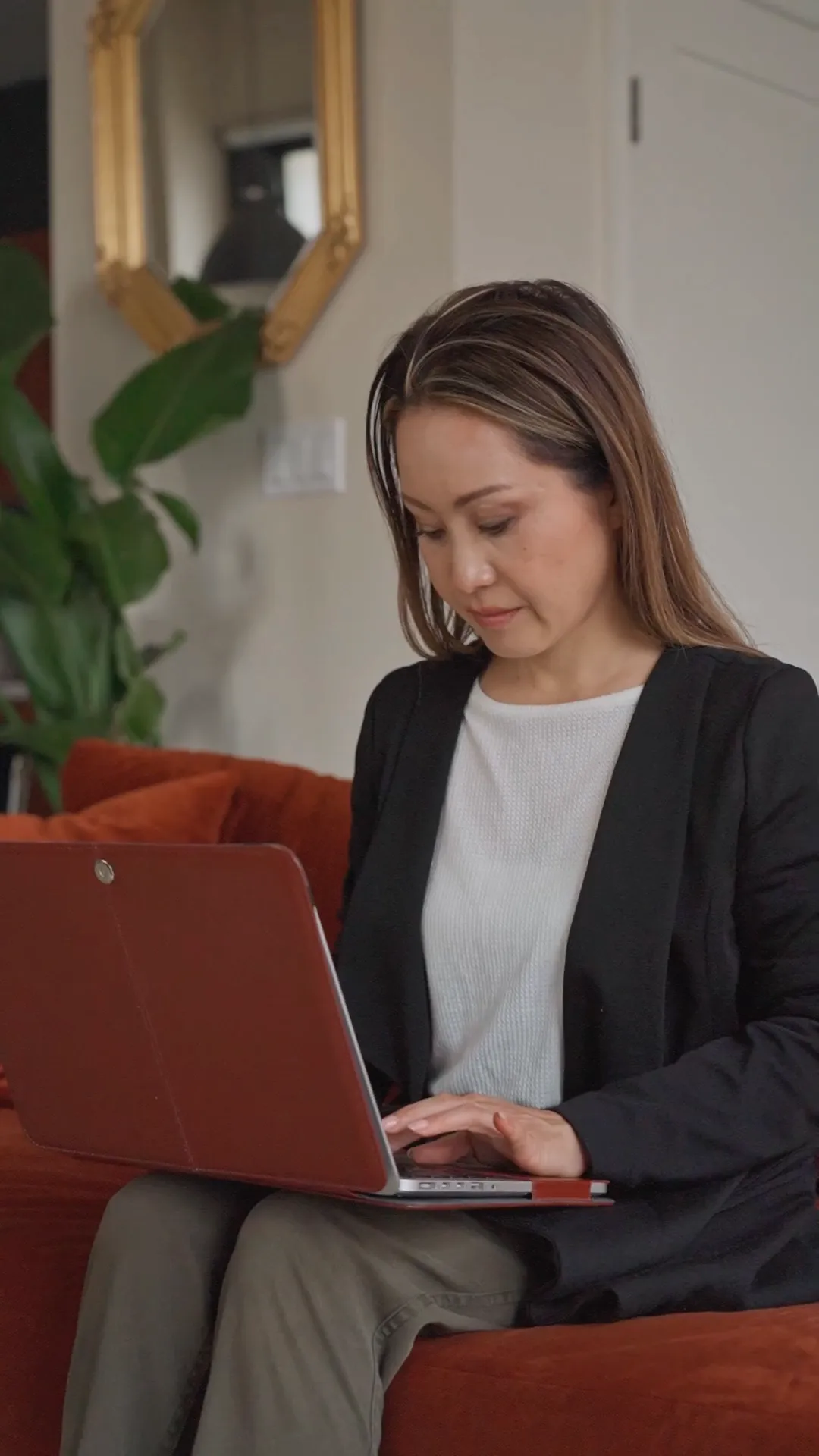 Woman sitting on a rust-colored couch using a laptop with a brown cover in a living room with green plants and a gold-framed mirror.