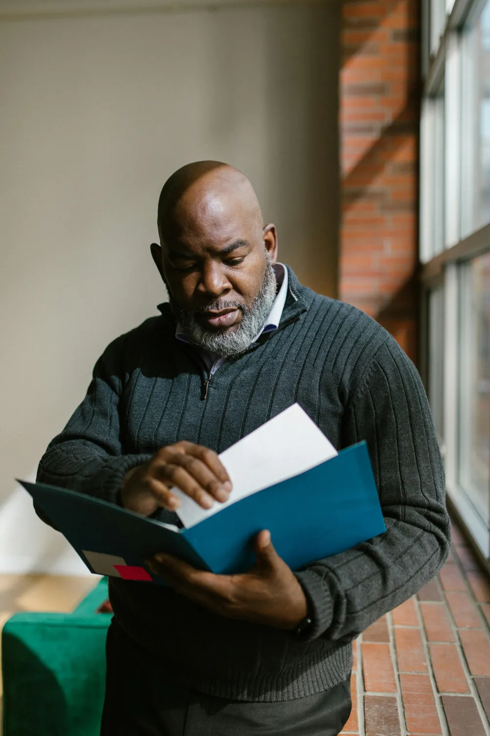 Mature man in a dark sweater reading documents in a blue folder near a window.