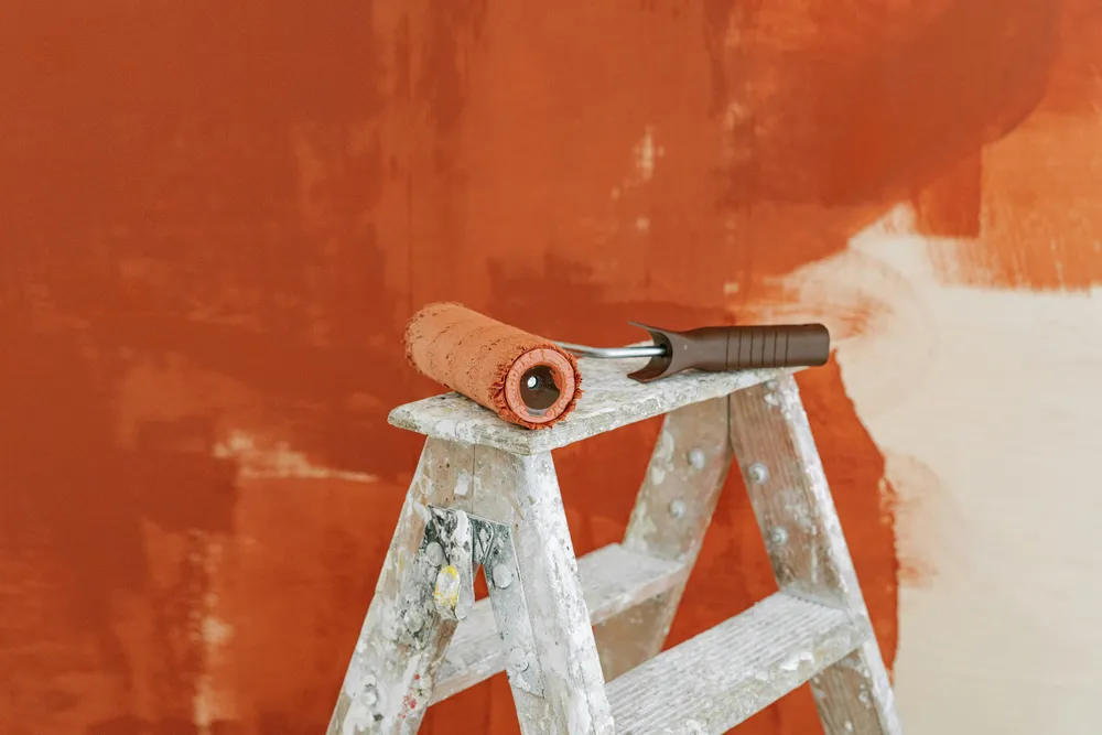 Paint roller covered in orange paint resting on a paint-splattered wooden ladder in front of a partially painted orange wall.