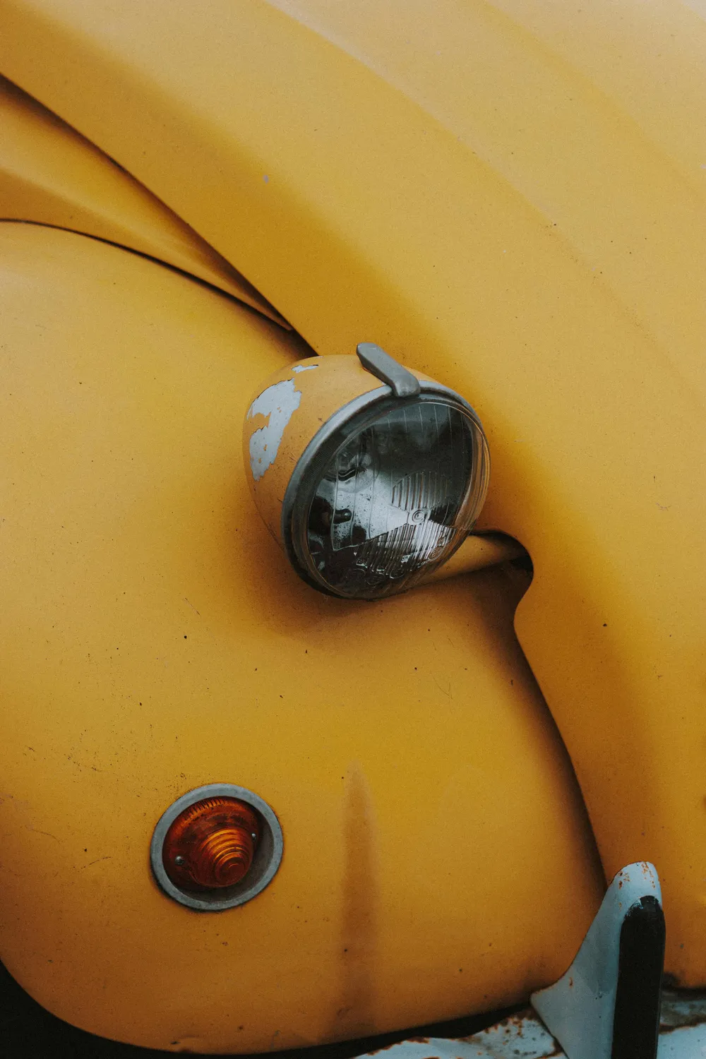 Close-up of a yellow vintage car's headlight and orange signal light with chipped paint and rust spots.