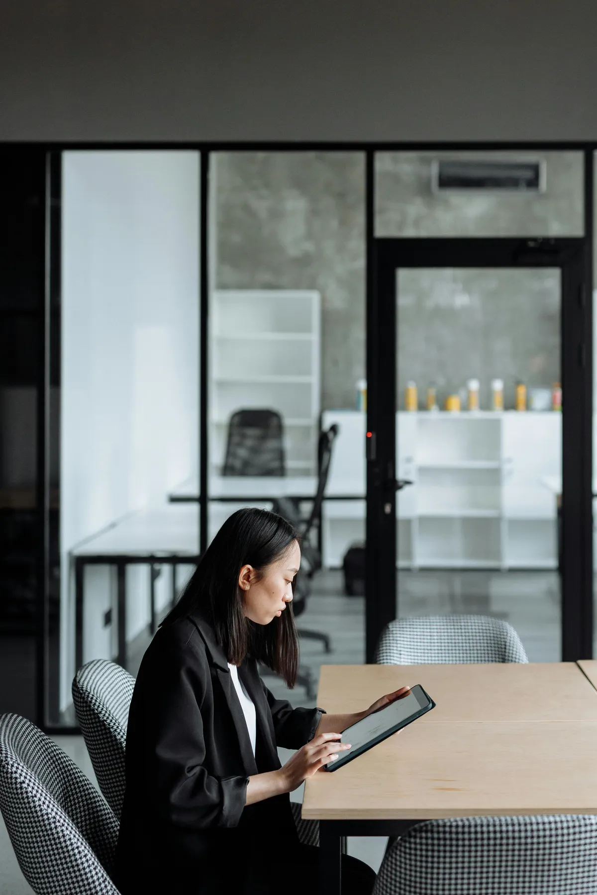 Woman in black blazer sitting at a table using a digital tablet in a modern office with glass walls.