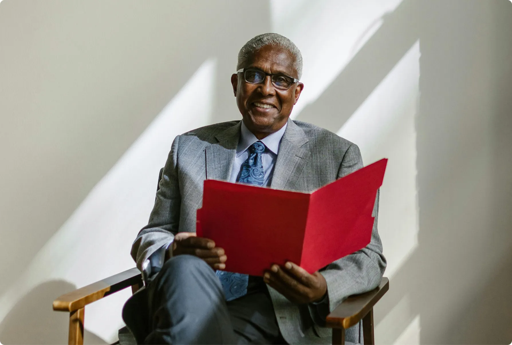 Smiling elderly man wearing glasses and a gray suit sitting on a wooden chair holding a red folder.