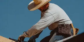 Construction worker in a hat and gloves working on a wooden roof under a clear blue sky.