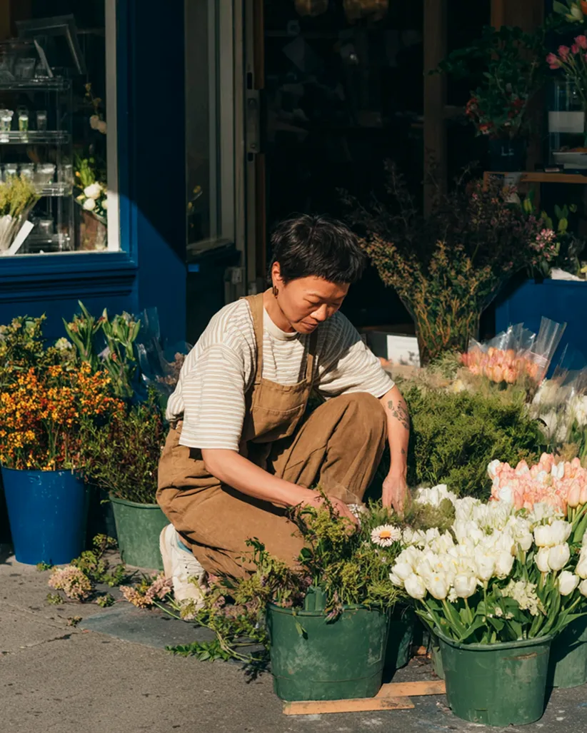 Person crouching outdoors arranging flowers in green pots outside a flower shop with various blooms.