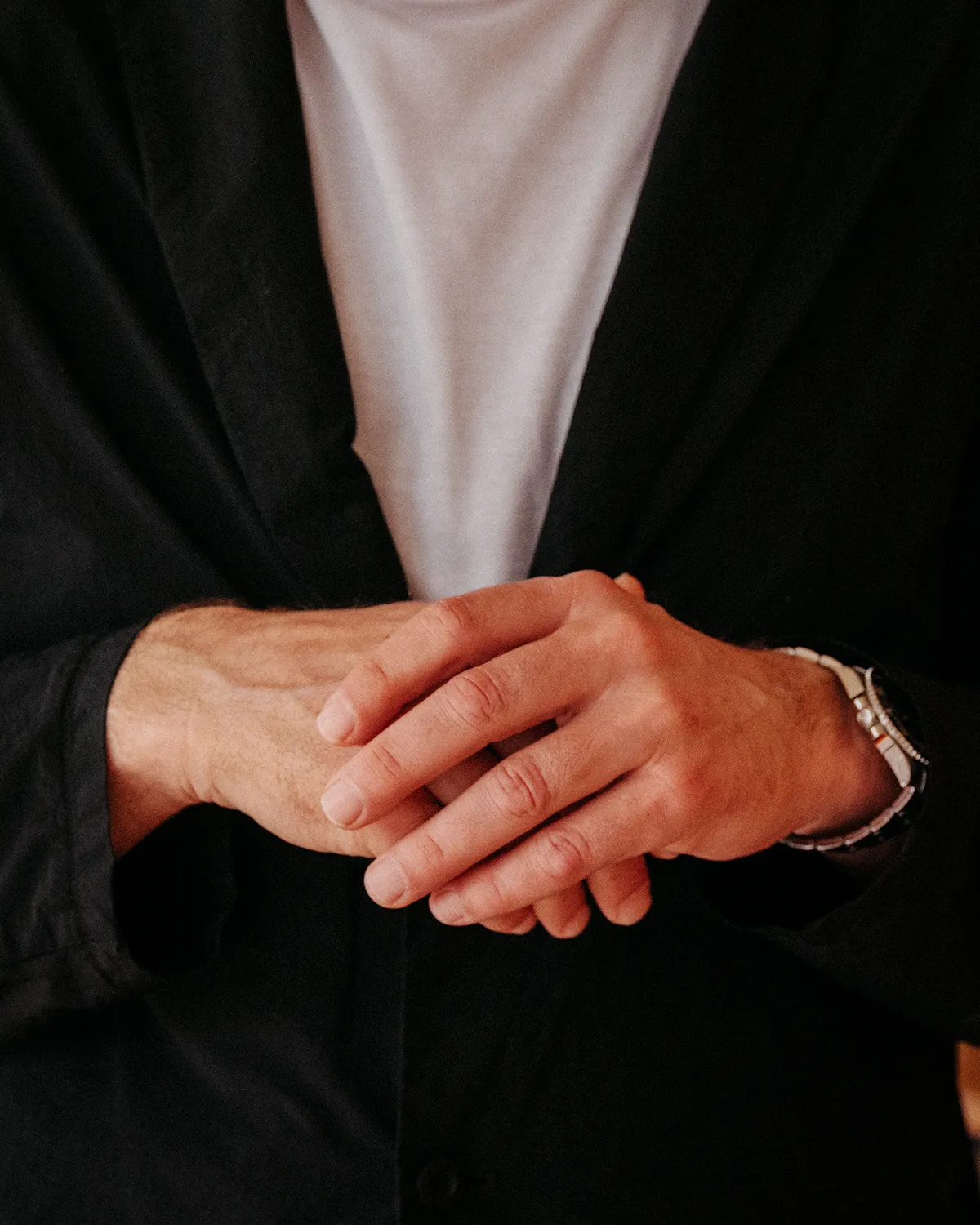 Man's hands clasped together wearing a black jacket, white shirt, and wristwatch.