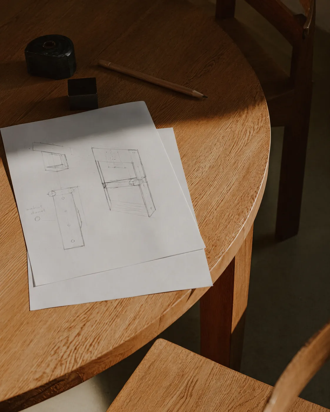 Wooden table with two sheets of paper showing furniture sketches, a pencil, and two small black objects.
