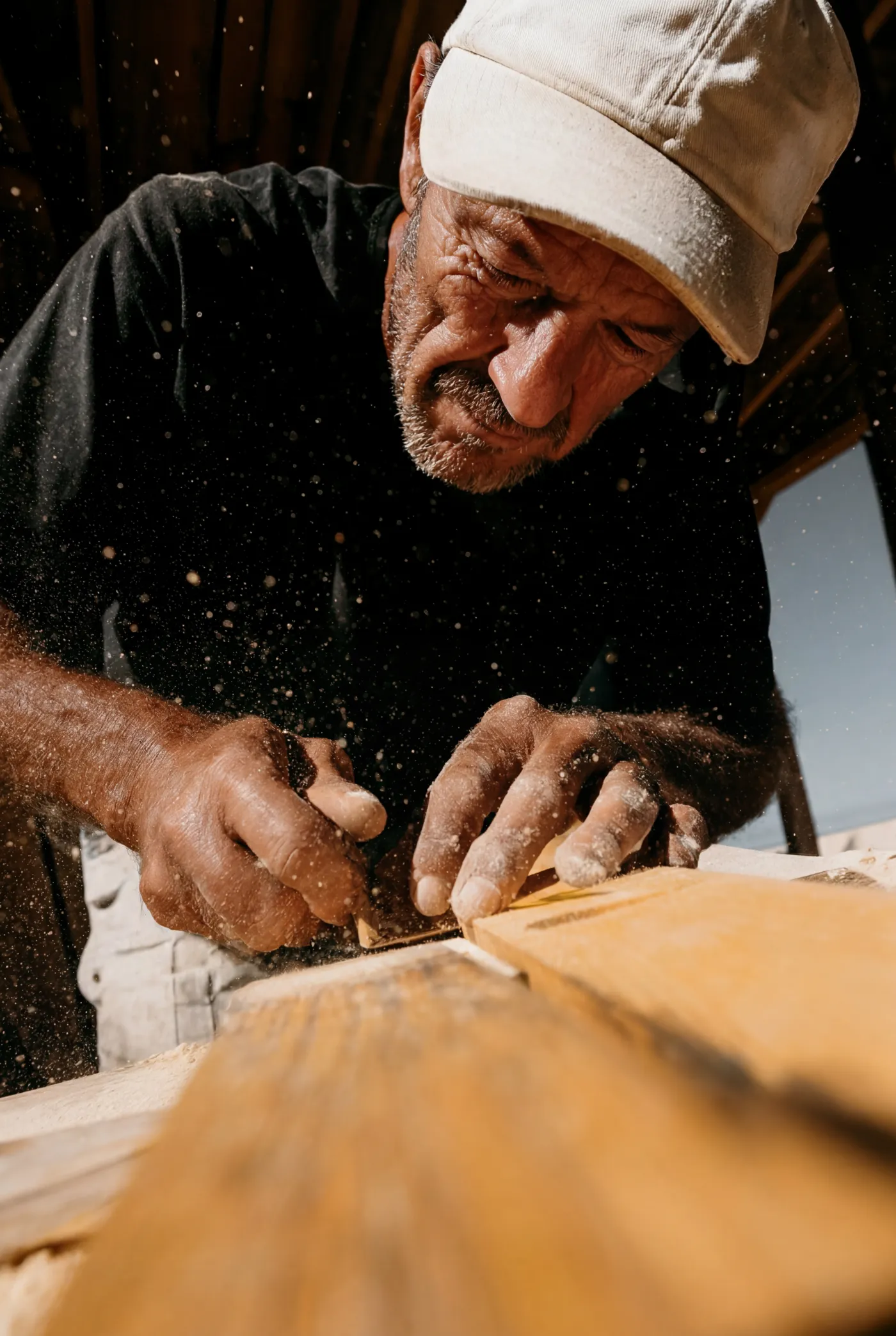 Man wearing a cap carving wood with intense focus while wood shavings fly around him.
