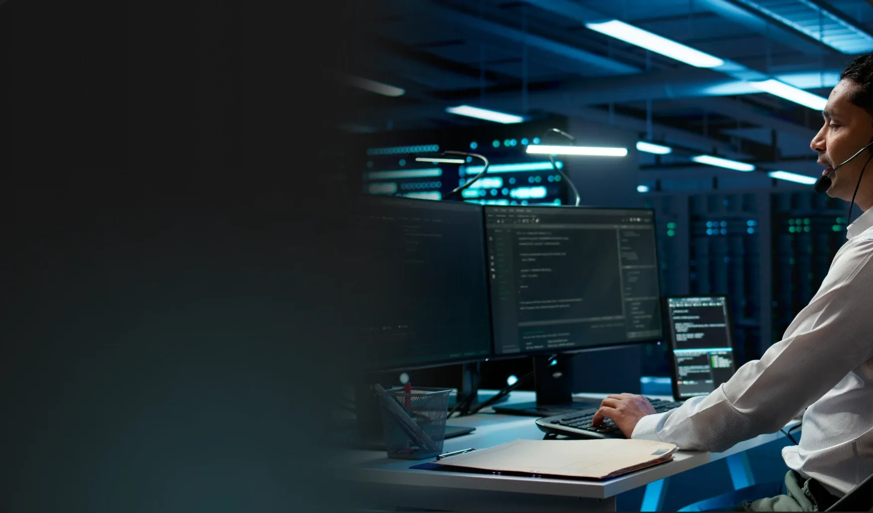 Hombre con auriculares y micrófono trabajando en una computadora con múltiples pantallas en un centro de datos moderno con iluminación azul.