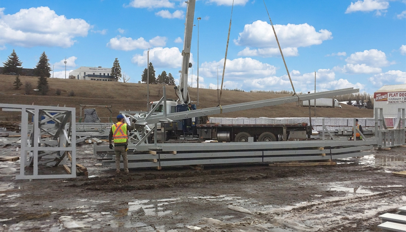 Workers in safety vests guiding a crane lifting metal beams at a muddy outdoor construction site.