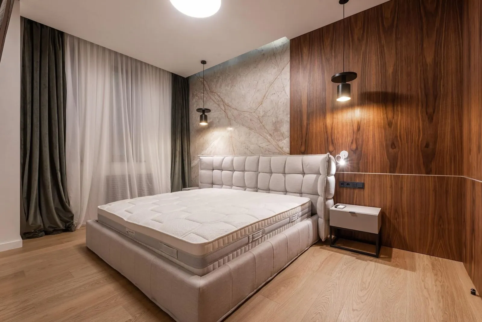 Modern bedroom with a light beige upholstered bed, marble and wood accent walls, pendant lights, and sheer curtains.