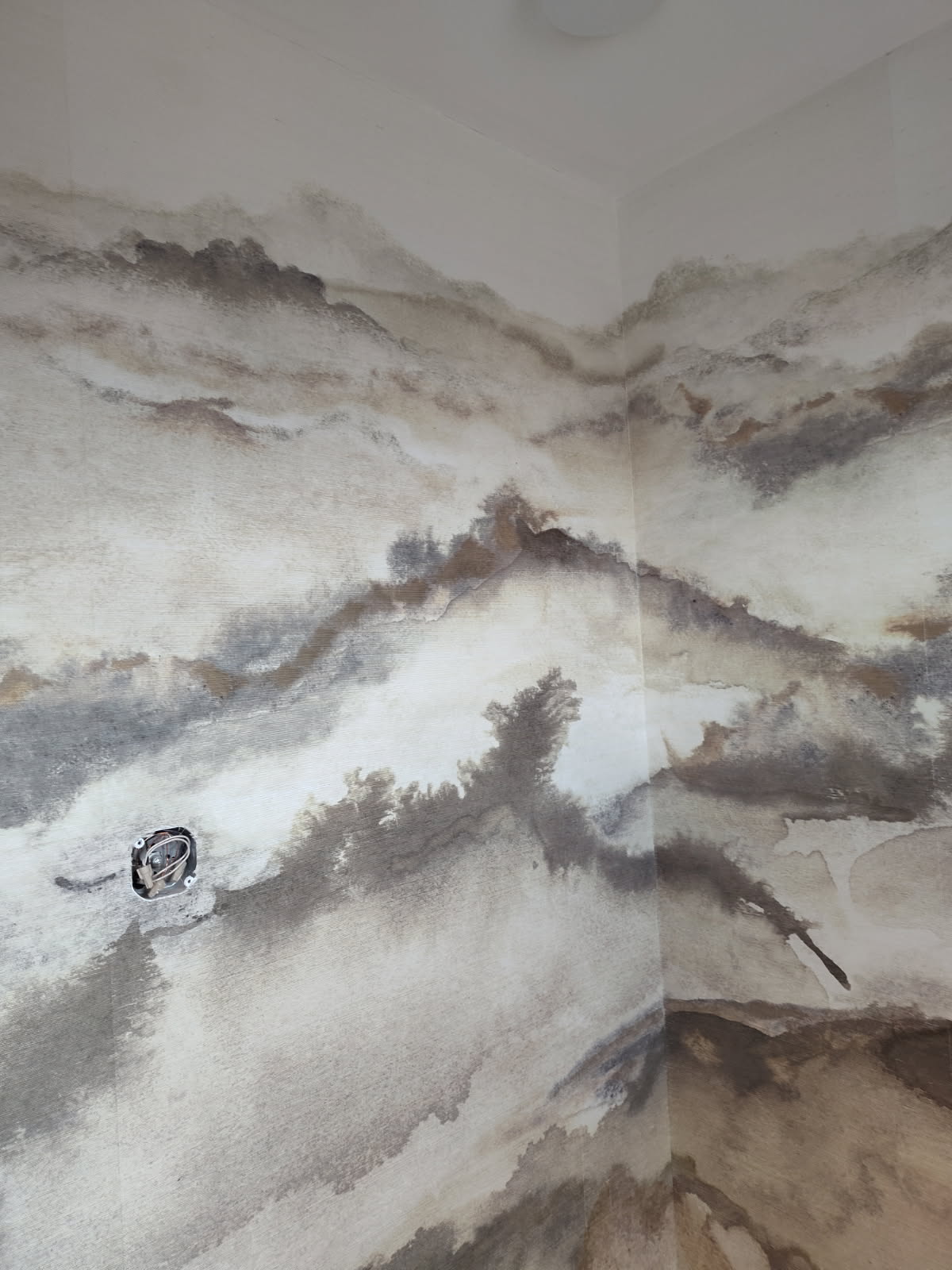 Corner of a wall covered with abstract gray and brown textured wallpaper with exposed wiring hole.