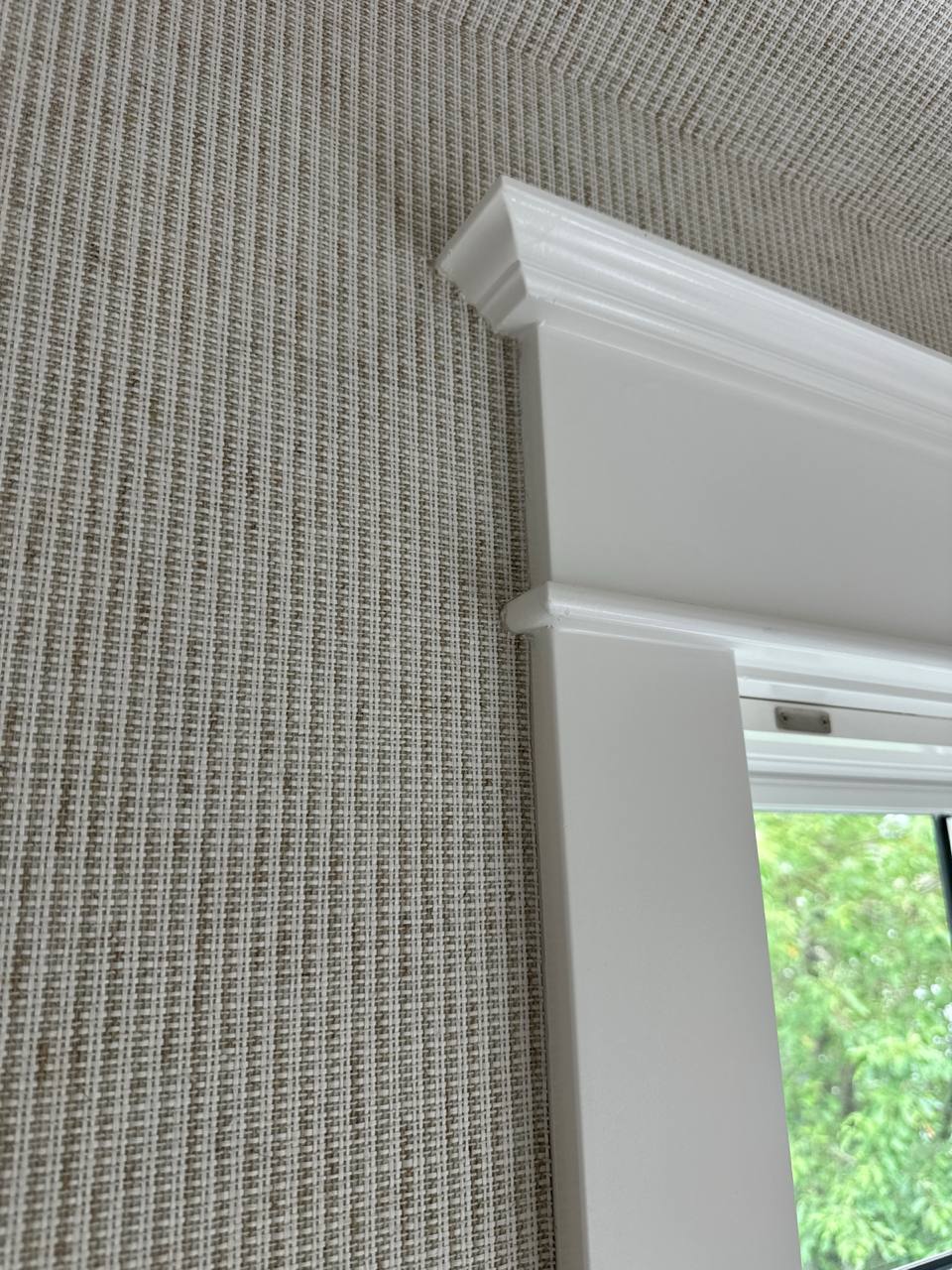 Corner of a white window frame with decorative molding against textured grayish wallpaper.