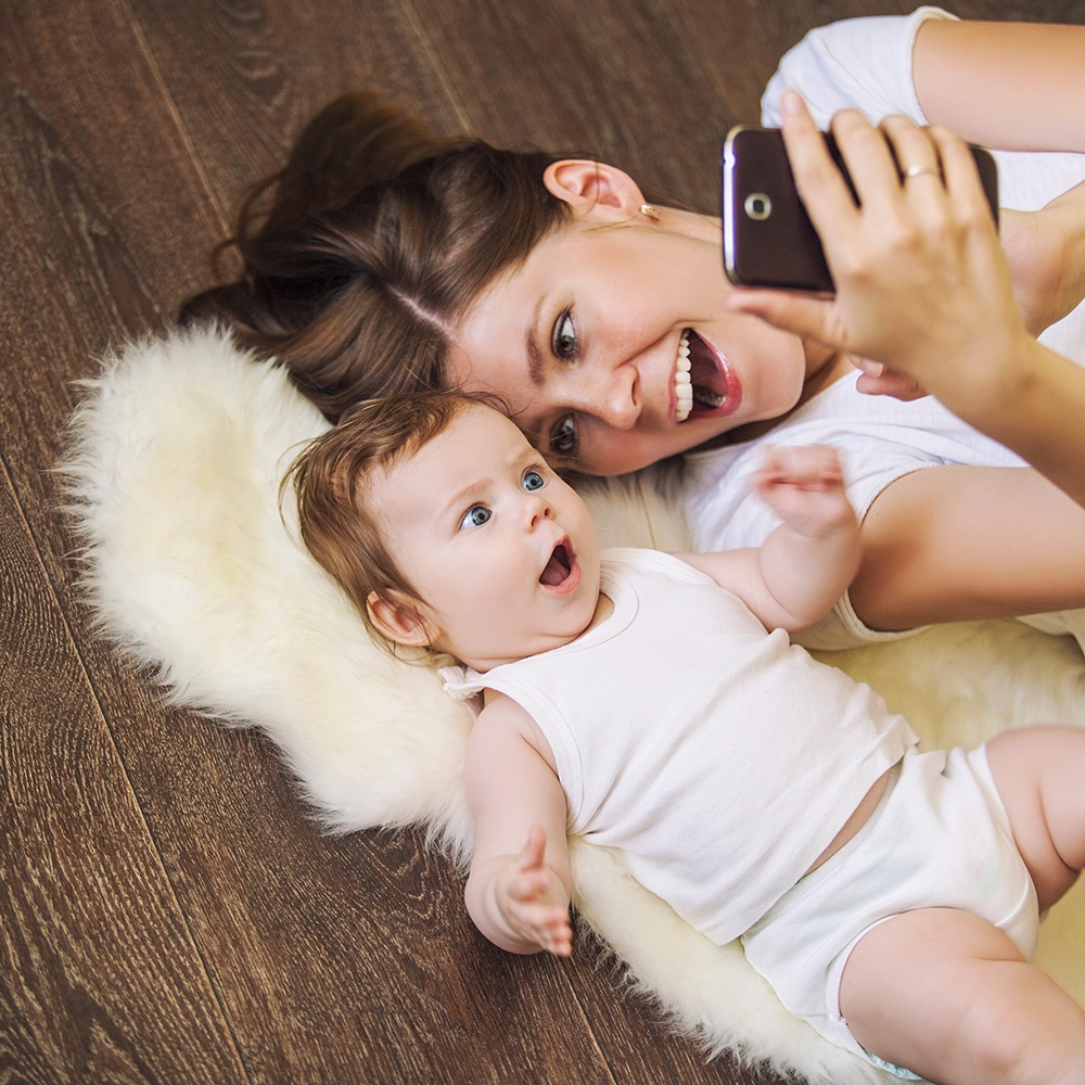 Mom and Baby lay on hardwood flooring taking a selfie with cell phone.