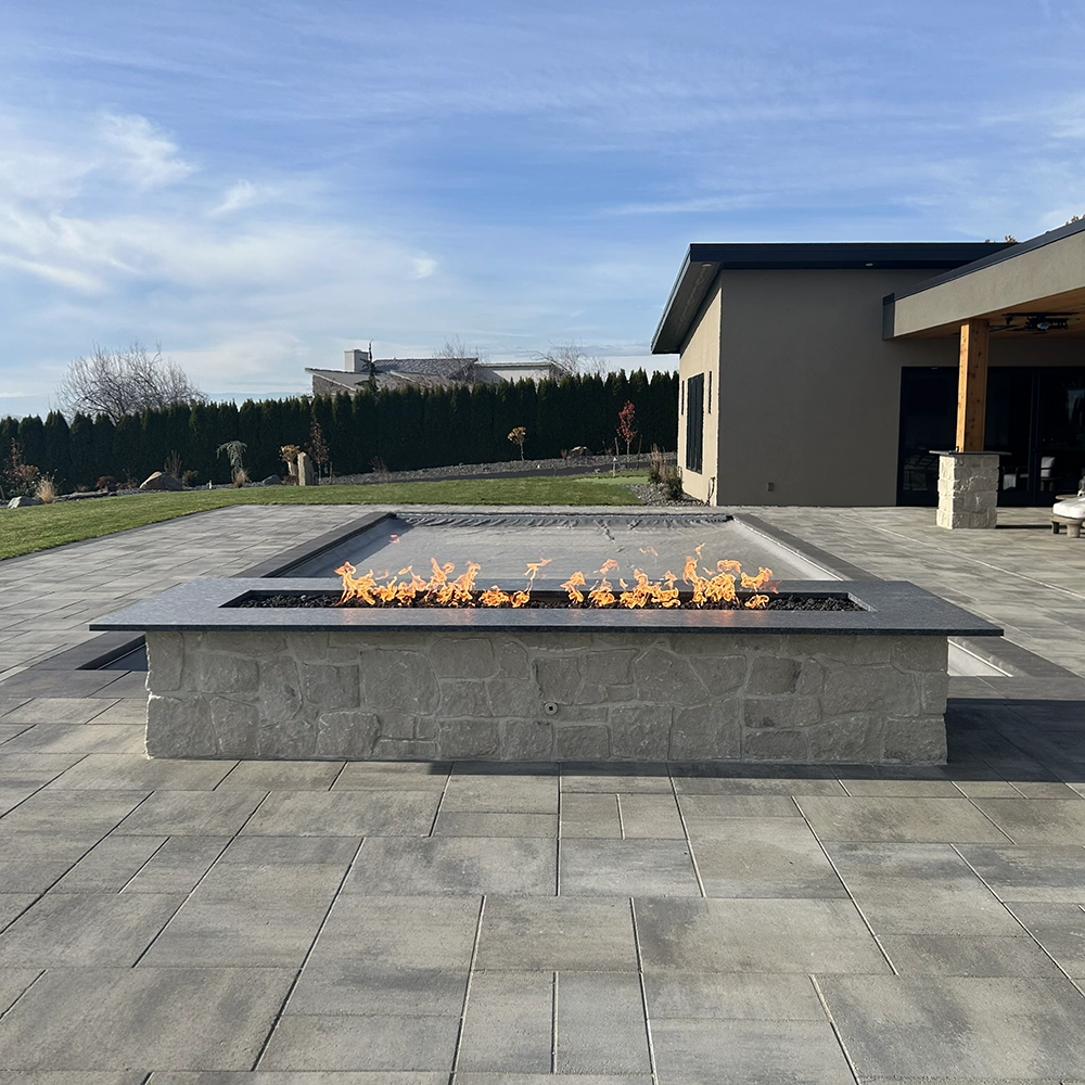 Rectangular granite fire pit with flames on a stone patio beside a lawn, pool, and modern house.