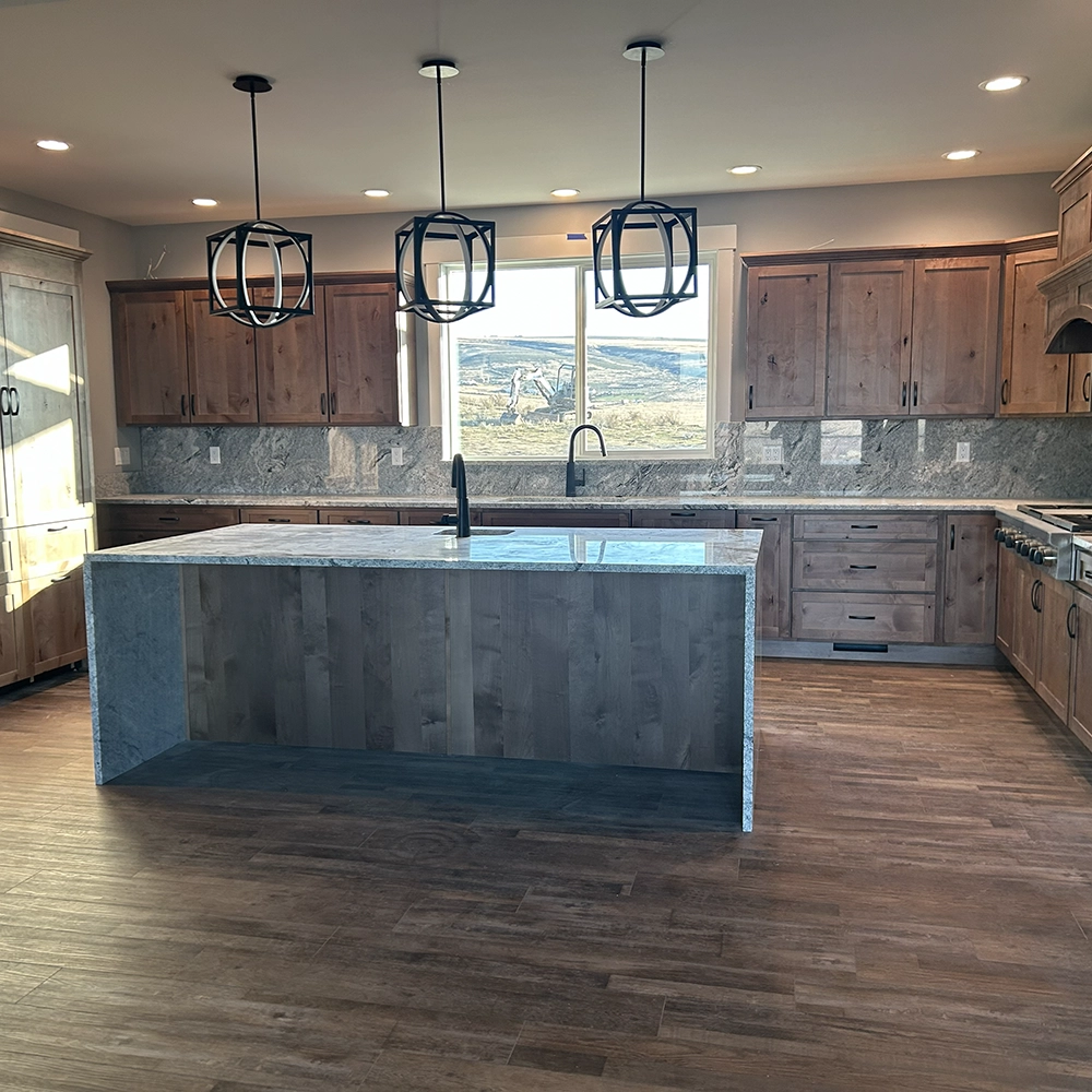 Large granite island with wood cabinets and three hanging pendant lights in a remodeled kitchen.