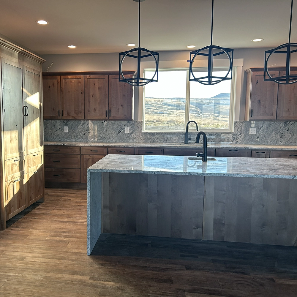 Granite island with a sink, wood cabinets, and geometric pendant lights below a window with a landscape view.