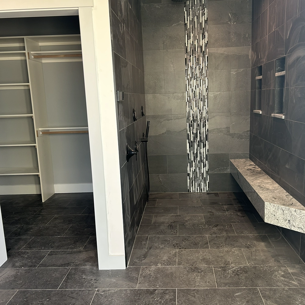Walk-in shower with dark gray tile, a vertical mosaic accent, and a built-in stone bench beside a closet.