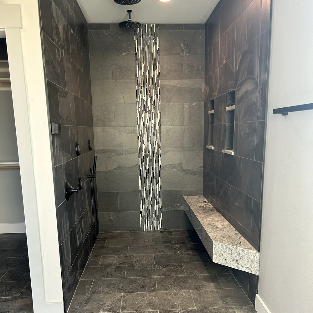 Walk-in shower with dark gray tile, a vertical mosaic strip, built-in niche shelves, and a stone bench.