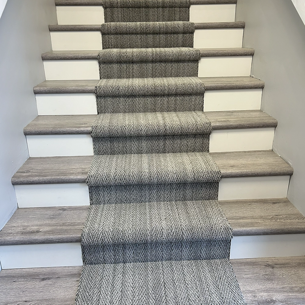 Wooden staircase with a light gray carpet runner featuring a subtle herringbone pattern down the center.