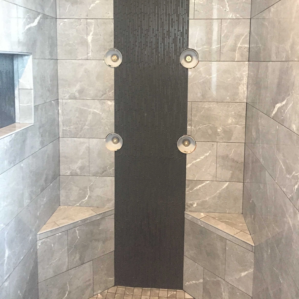 Gray tiled shower with a vertical strip of smaller dark tiles, four showerheads, and built-in corner seats.