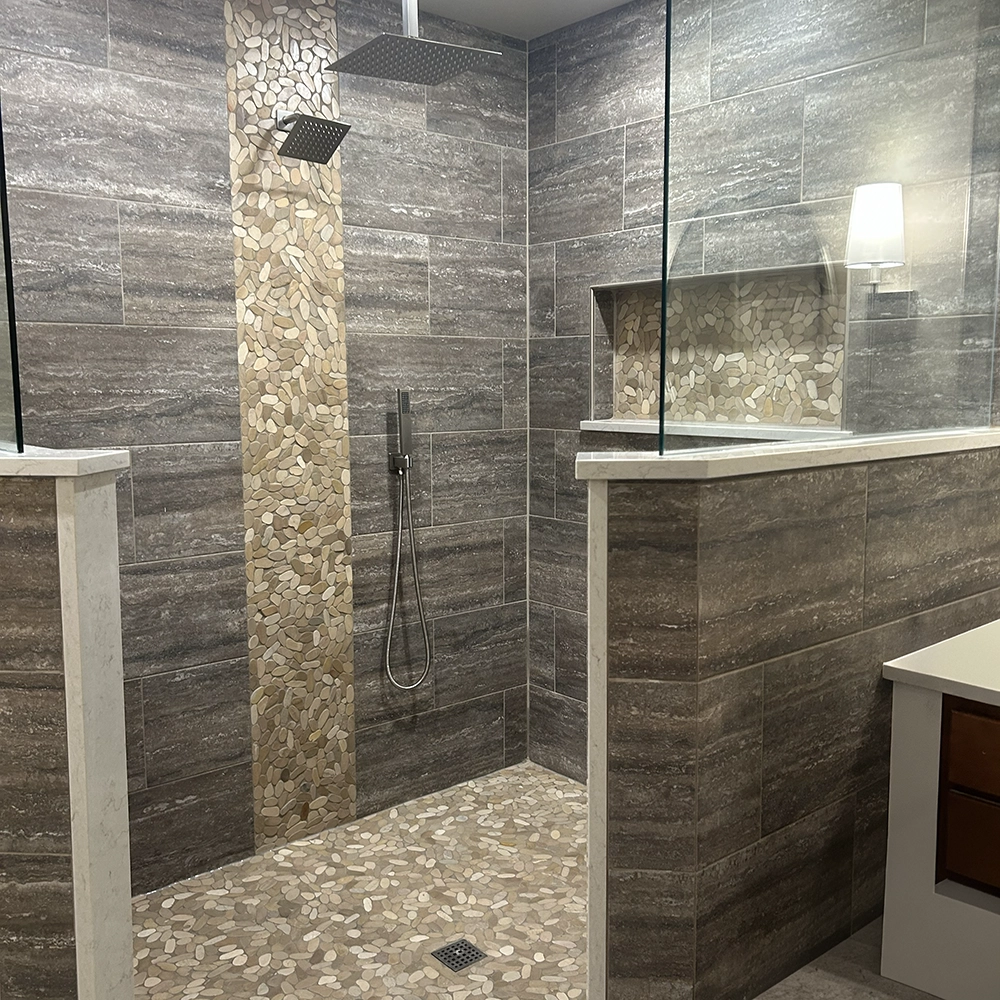 Walk-in shower with a pebble stone floor and accent strip, dark gray tile walls, and a square rain showerhead.