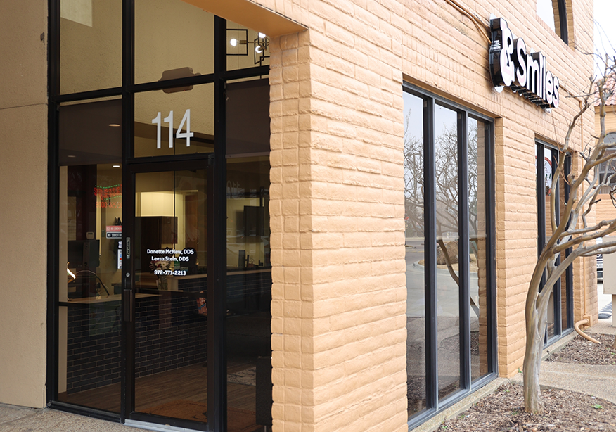 Exterior view of a dental office entrance with large glass doors and windows, showing the suite number 114 and signage for the practice.