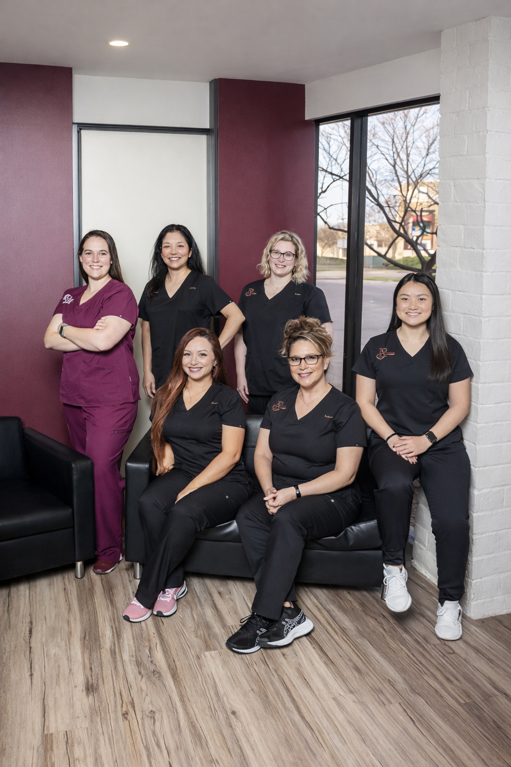 Group portrait of six dental team members in black and burgundy scrubs posing in a modern office lobby with seating and a window in the background.