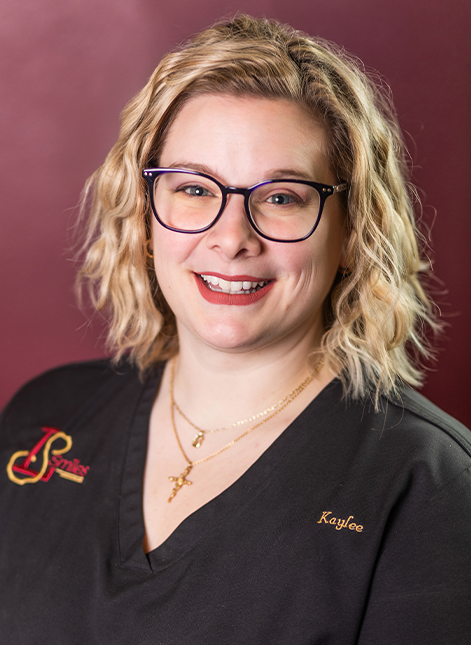 Portrait of Kaylee smiling in black scrubs against a burgundy background.