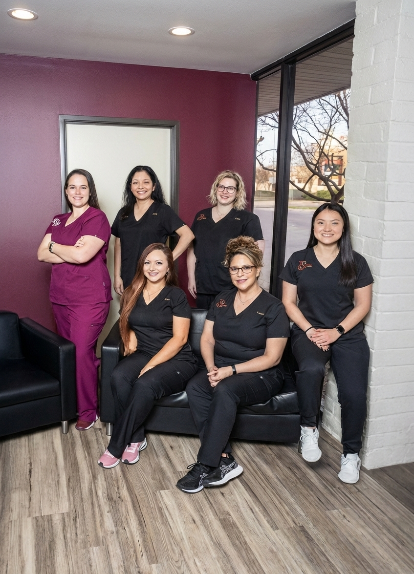 A portrait-oriented group photo of six smiling medical staff members posing in an office waiting area. On the far left, a woman stands with arms crossed wearing maroon scrubs, while the other five women wear matching black scrubs. Two women are seated on a black leather sofa in the center, three stand behind them, and one leans against a white painted brick pillar on the right. The room features light wood laminate flooring, deep maroon walls, and large windows in the background. The image has been vertically expanded to show the white ceiling with recessed lighting above and more of the wooden floor below.