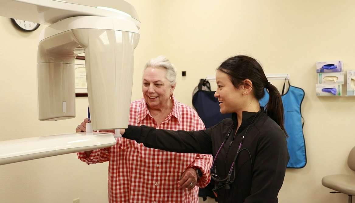A smiling dental assistant and an older woman are next to a dental imaging machine. The image is extended to a landscape format, showing more of the room.