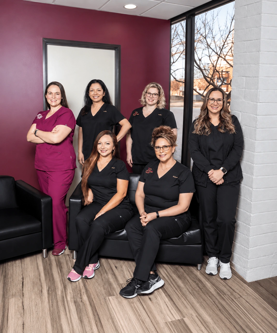 Five smiling dental team members pose together in a modern dental office reception area with burgundy accent walls, black leather seating, and large windows. Three staff members stand behind two seated colleagues, all wearing professional scrubs, creating a warm and welcoming group portrait in a bright, contemporary clinic.