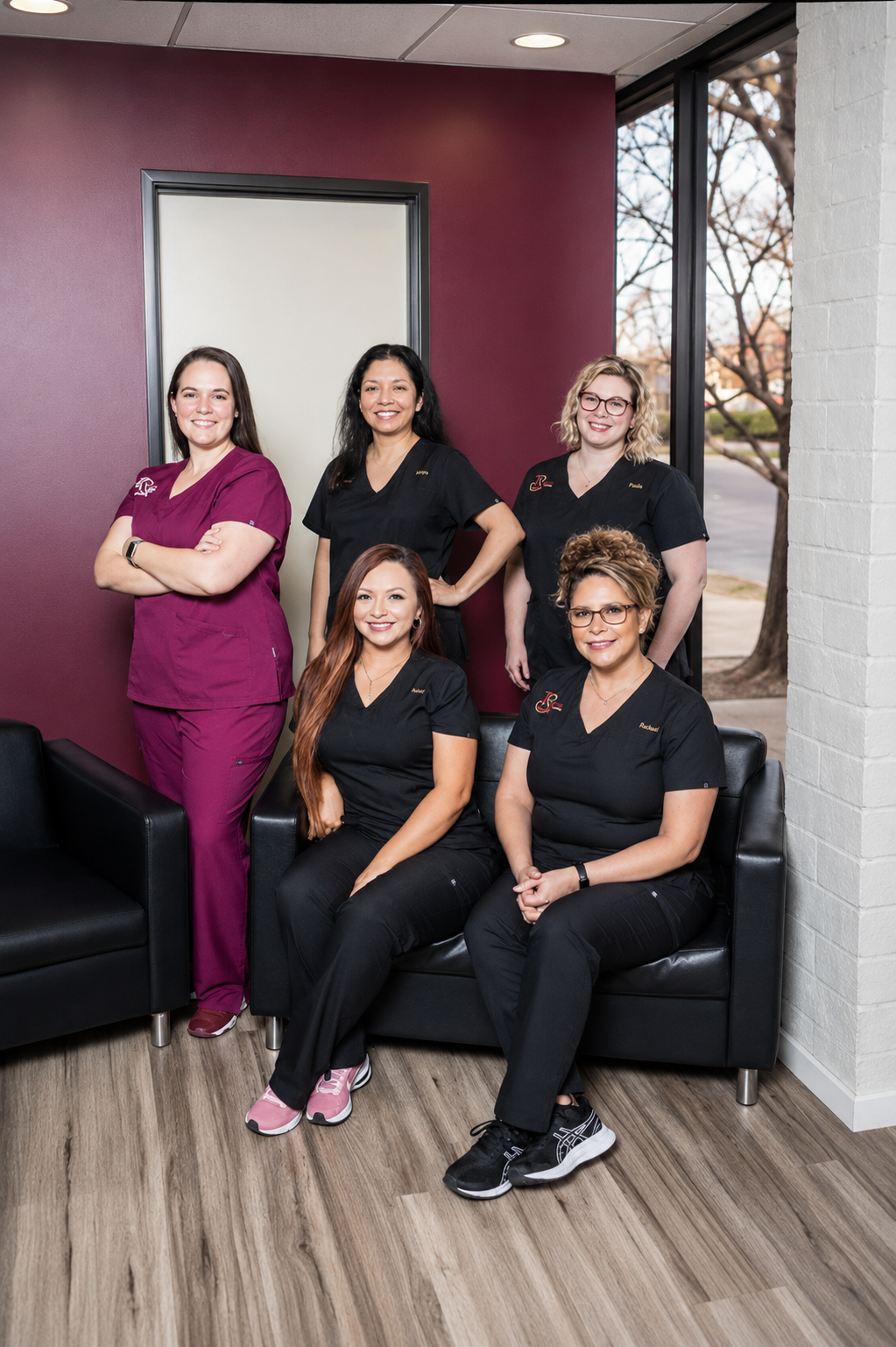 Five smiling dental team members pose together in a modern dental office reception area with burgundy accent walls, black leather seating, and large windows. Three staff members stand behind two seated colleagues, all wearing professional scrubs, creating a warm and welcoming group portrait in a bright, contemporary clinic.