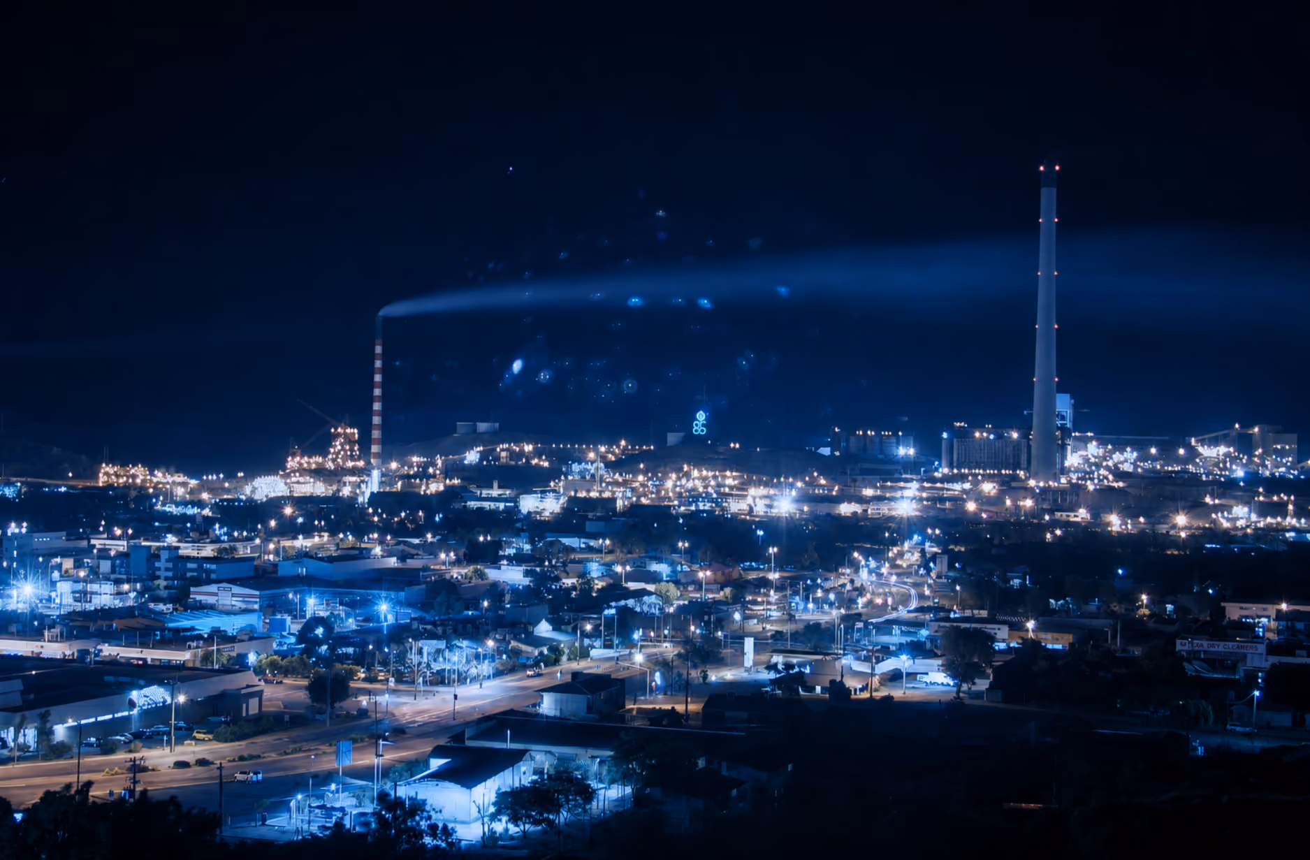Industrial area at night with bright lights and smoke coming from a striped smokestack.