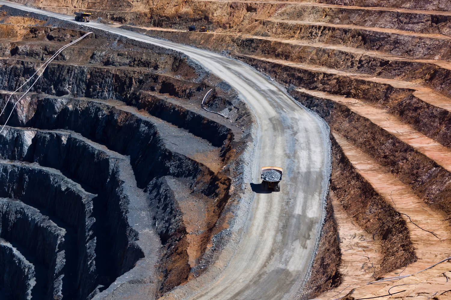 Large mining truck carrying rocks on winding road in an open-pit mine with terraced earth layers.
