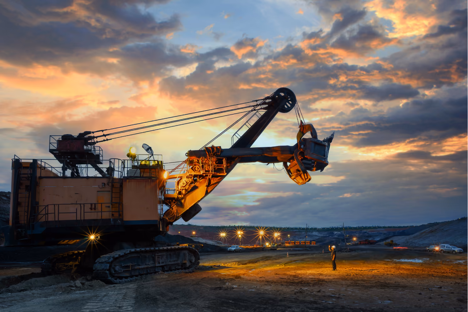 Large mining excavator with bucket raised, worker standing nearby under a dramatic sunset sky.