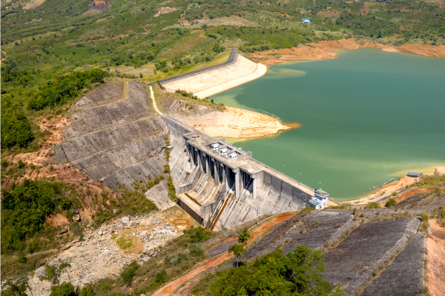 Concrete dam holding a large reservoir surrounded by green hills and rocky terrain.