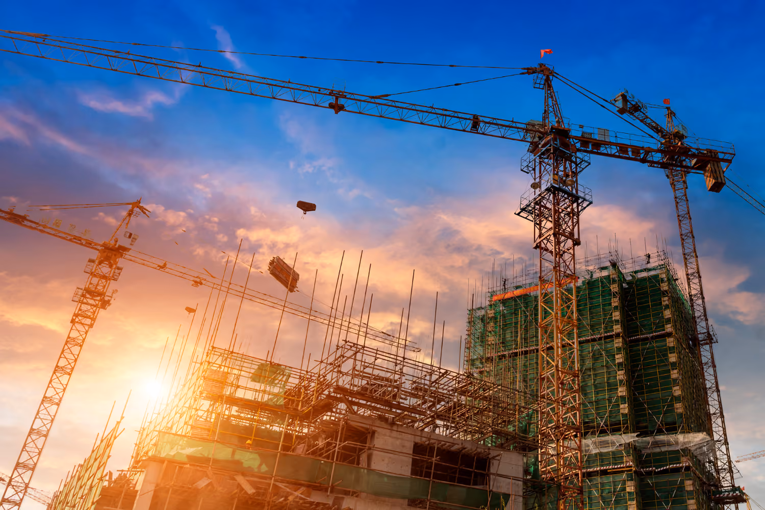 Construction site with cranes and scaffolding around buildings under a sunset sky.