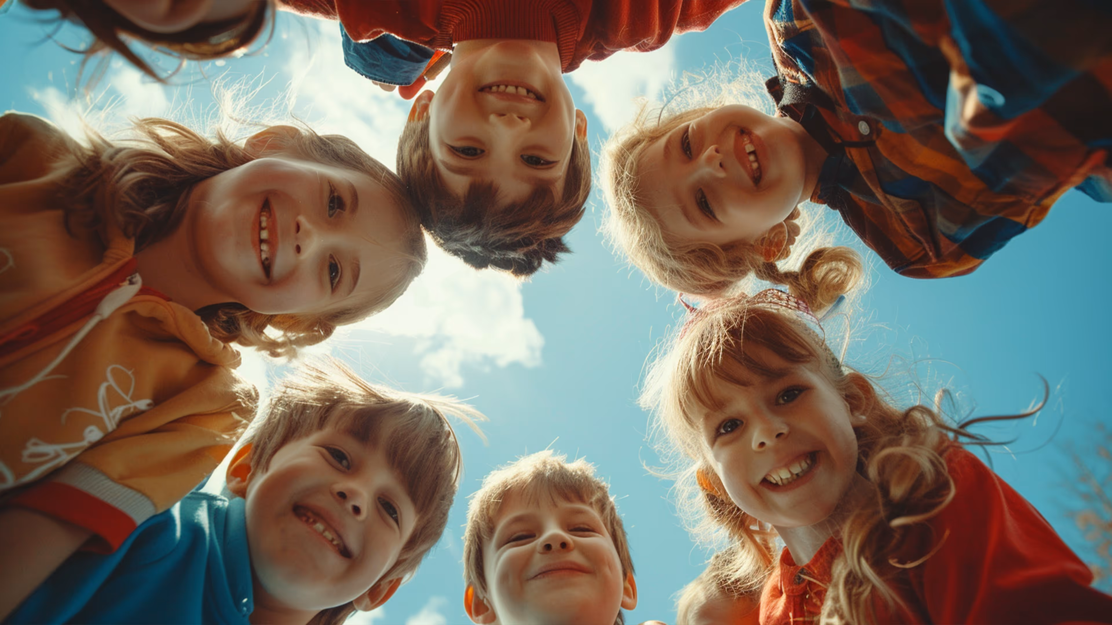 Group of seven smiling children in colorful clothes standing in a circle outdoors looking down at the camera against a blue sky with clouds.